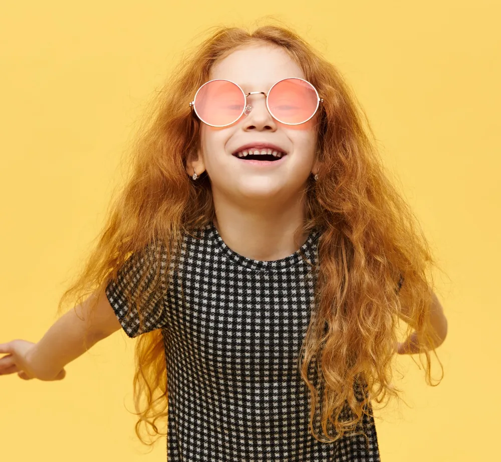 A laughing girl with long red hair and pink glasses against a yellow background